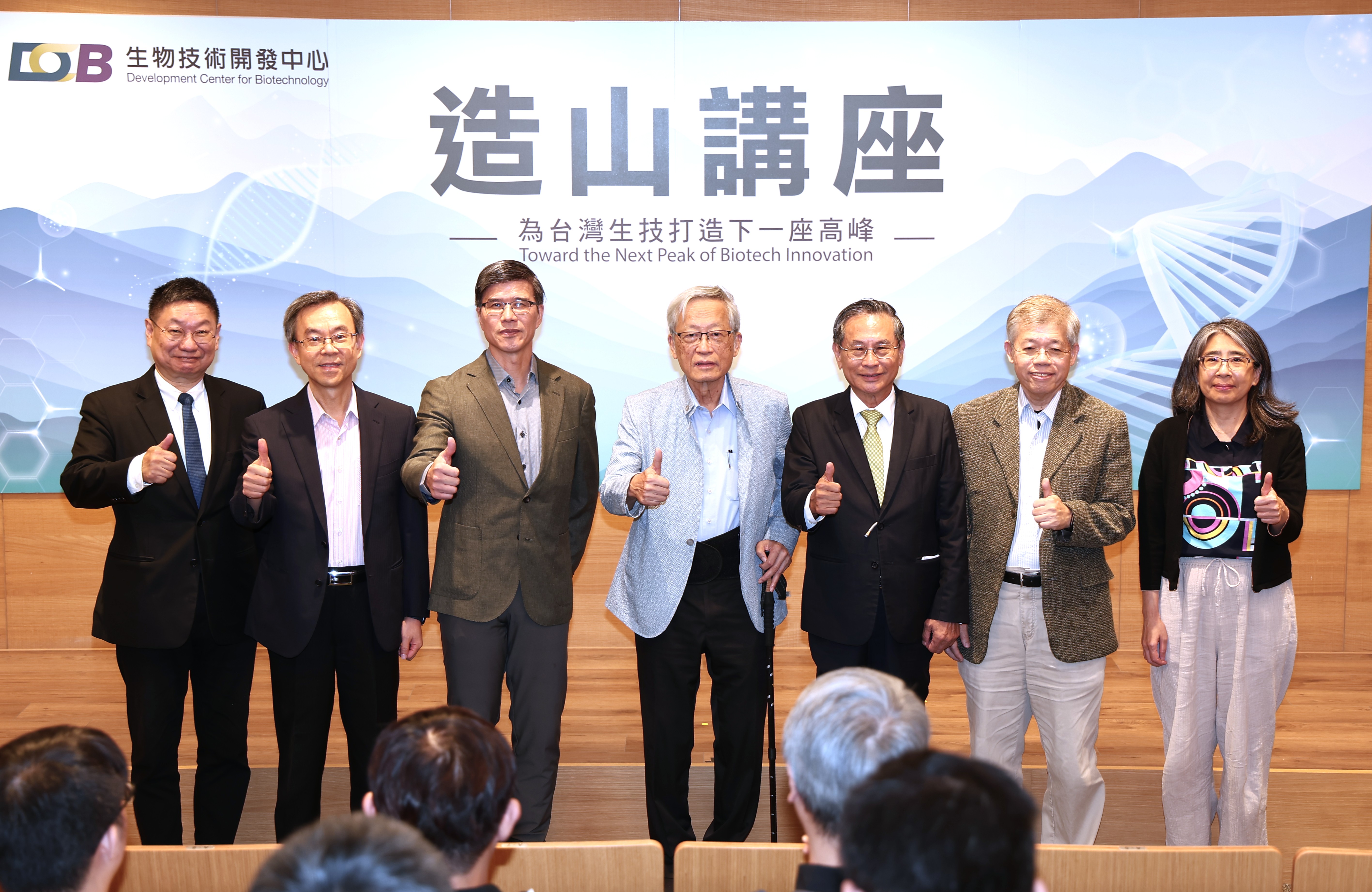 照片呈現一場生技相關活動或正式發表會，多位男女貴賓站在舞台前合影留念。背景看板以藍白色幾何圖案、科技光線與分子結構為設計元素，並寫有「生物技術開發中心」、「邁向生技創新高峰」等標語。畫面上方可見中心標誌及英文名稱，現場氣氛莊重而充滿未來感。參與者大多穿著西裝、外套或正式服裝，其中幾人面帶微笑並豎起大拇指，展現合作成功與慶祝活動成果的喜悅。前景可看到觀眾的頭部，顯示這是一場面向群眾或業界人士的公開場合。